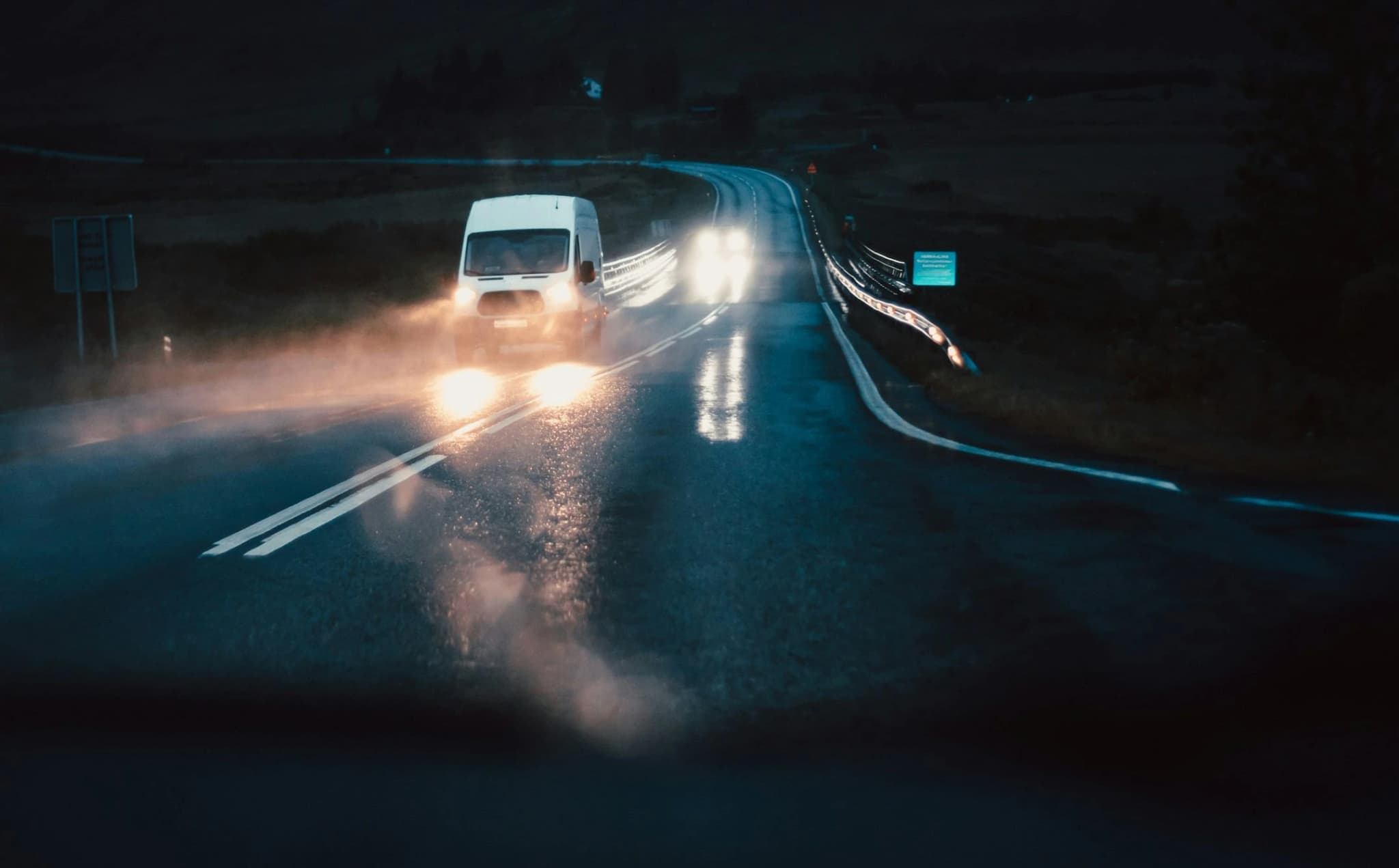 A light commercial van with its headlights on, driving a wet road at night